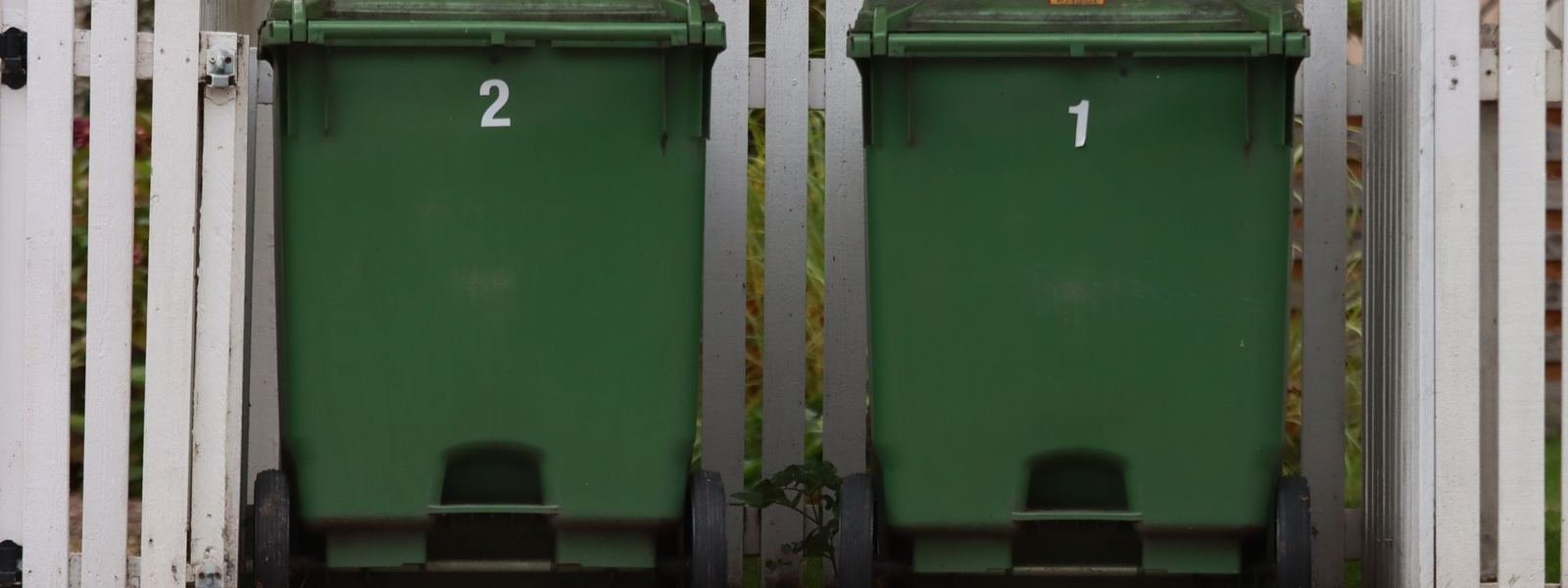 Two green wheelie bins beside a white fence
